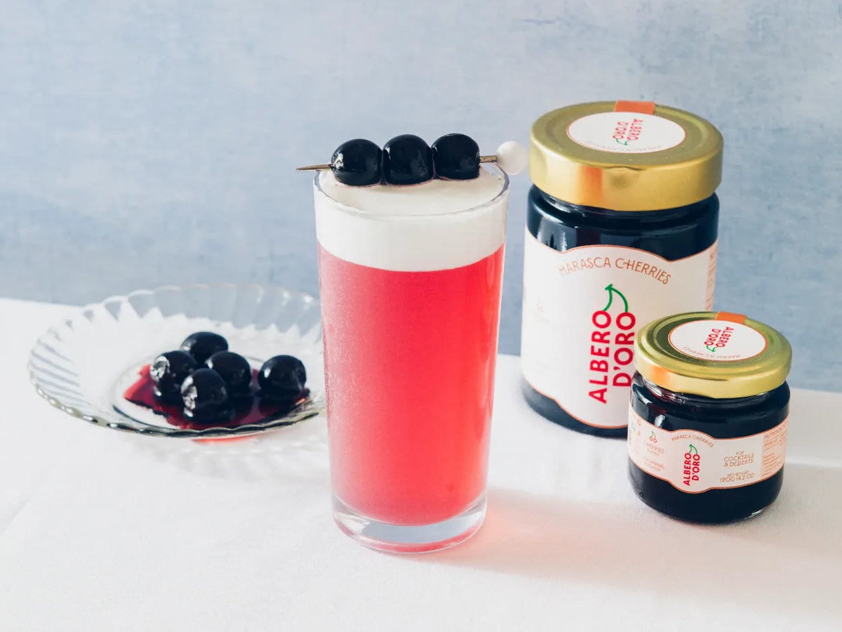 Pink cocktail garnished with a skewer of Albero d'Oro marasca cherries, next to two jars of marasca cherries and a dish of cherries